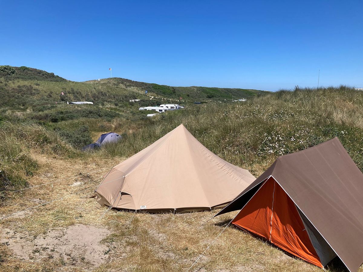 Zwei Zelte auf einer Dünenwiese unter blauem Himmel, dahinter Wohnwagen